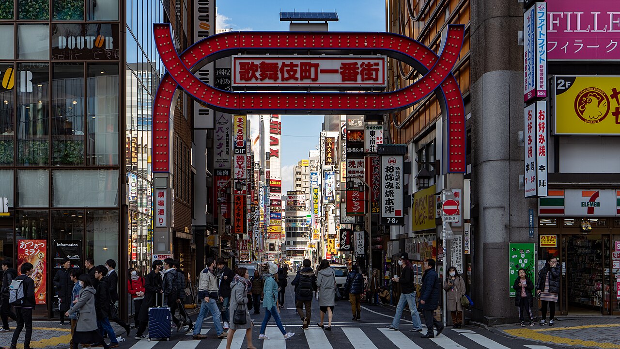 The red illuminated Kabukicho Ichibangai archway over a crowded Shinjuku street in daylight, with layered neon signage receding behind it