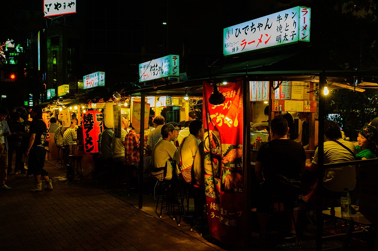 A row of lit yatai food stalls along the Naka River in Fukuoka at night, with customers seated under the awnings and office towers behind