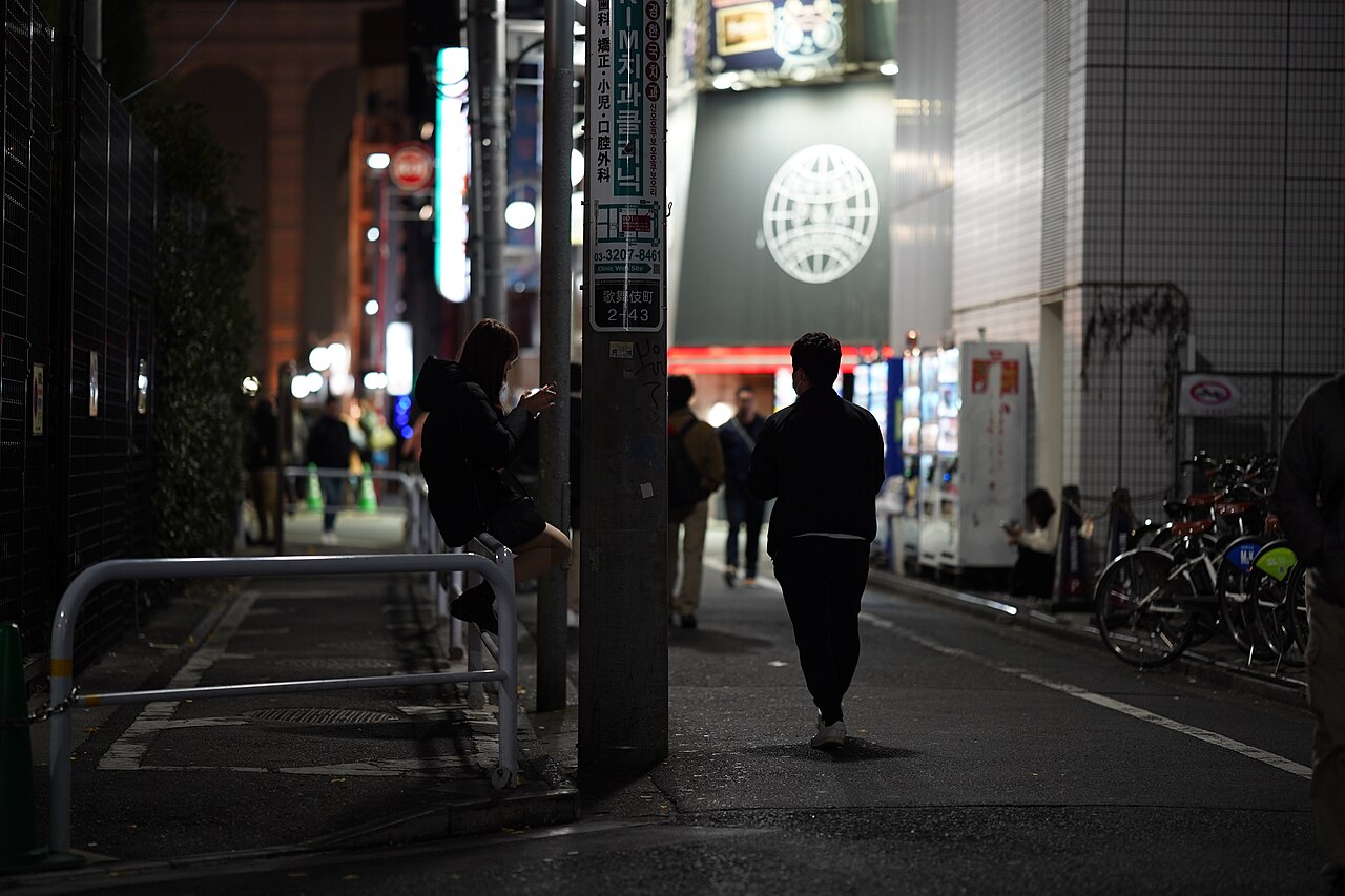 A dim Kabukicho side street at night near Okubo Park, with a lone figure seated on a guardrail and pedestrians passing under neon light