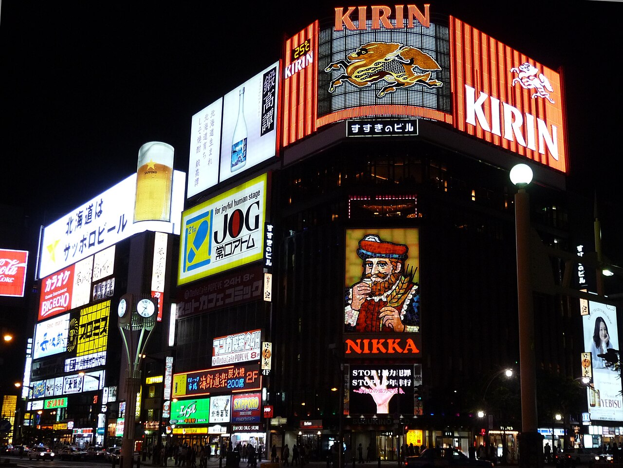 The Susukino crossing in Sapporo at night, dominated by large illuminated Kirin and Nikka signs above a wall of stacked neon storefront boards