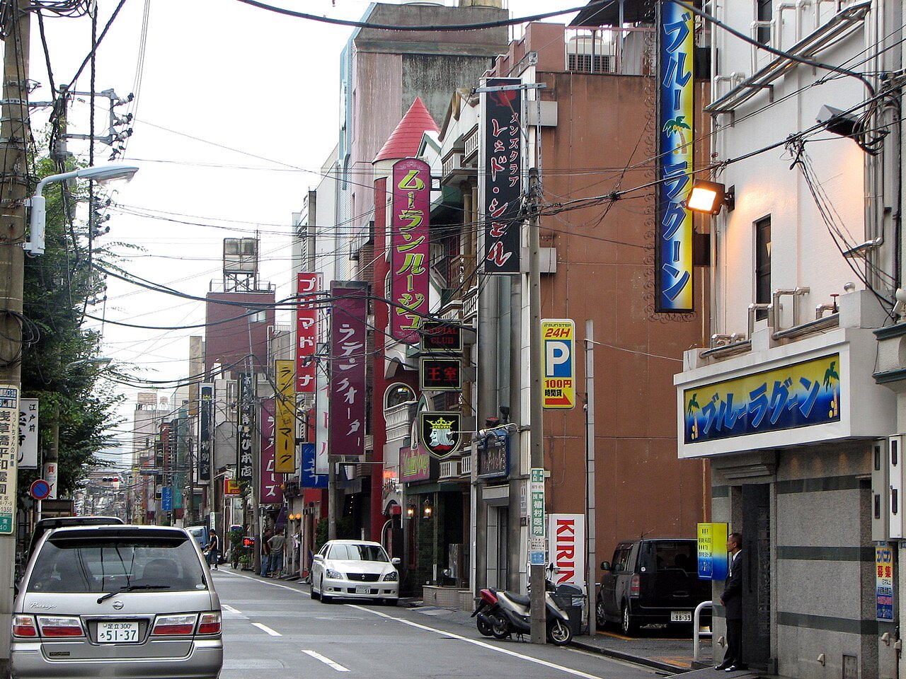 A narrow Yoshiwara street in Tokyo lined with vertical soapland signboards in Japanese, parked cars and a man in a dark suit standing at a shopfront